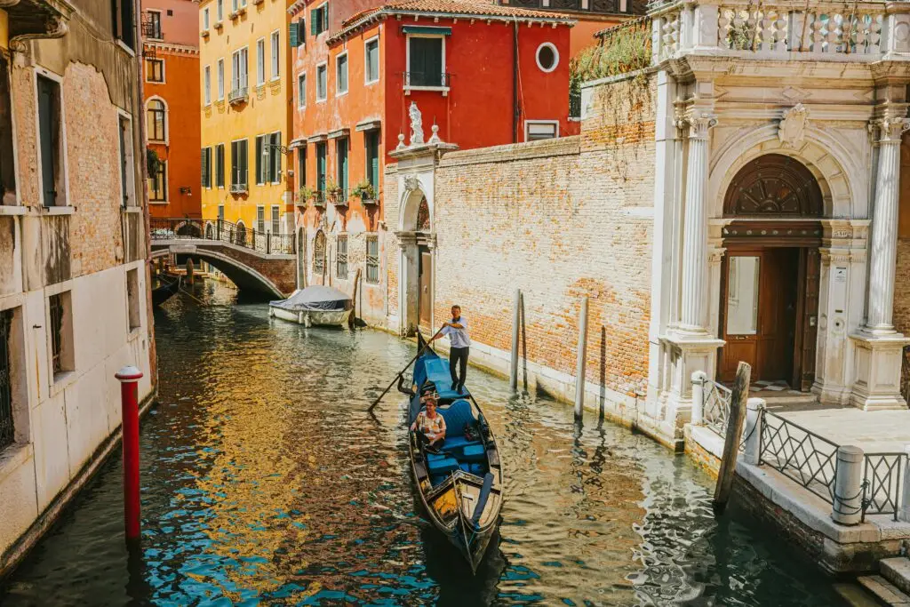 A traditional Venetian gondola with a gondolier navigating a narrow, sunlit canal lined with historic orange and yellow buildings