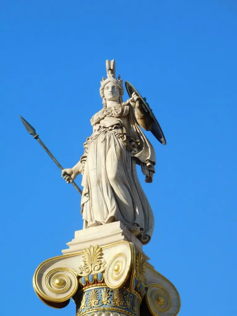 A majestic white marble statue of the Greek goddess Athena holding a spear and shield against a vibrant blue sky