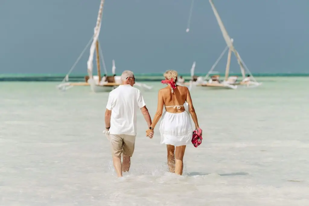 A couple walking hand-in-hand through crystal clear turquoise water toward traditional sailboats on a white sand beach.